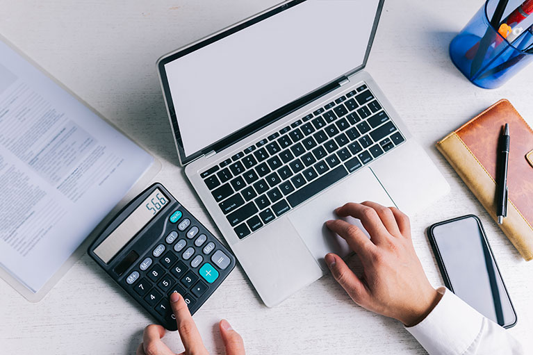 Photo d'un poste de travail avec la main d'une personne sur le clavier de son ordinateur et de l'autre main sur une calculatrice.