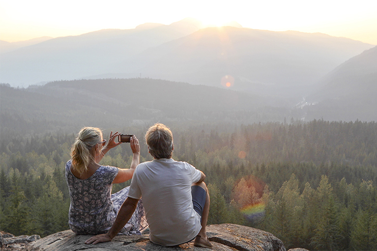 Ce qui nous distingue Alt text: image d’un homme et une femme assis, qui prennent une photo de la vue en haut d’une montagne au coucher du soleil. 