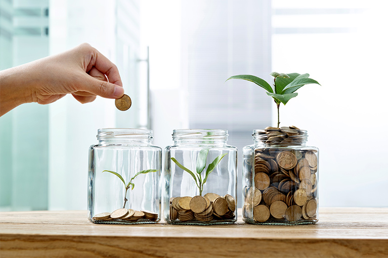 Trois verres placés sur une table dont deux contiennent de la monnaie et une plante pour démontrer l’évolution. Une main qui dépose de la monnaie dans le troisième verre.