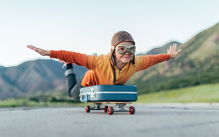 Un enfant avec un masque de pilote et les mains levé, couché sur une valise qui elle est installé sur un skateboard.