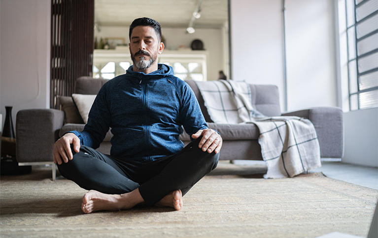 Un homme assis sur le tapis dans son salon en train de méditer.