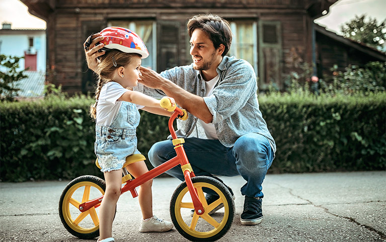 Un père à genou qui arrange le casque de son enfant assis sur un vélo.