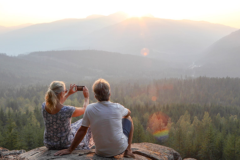 Couple de retraités assis sur une colline en forêt, la femme prenant une photo d’un paysage ensoleillé rempli de sapins verts.