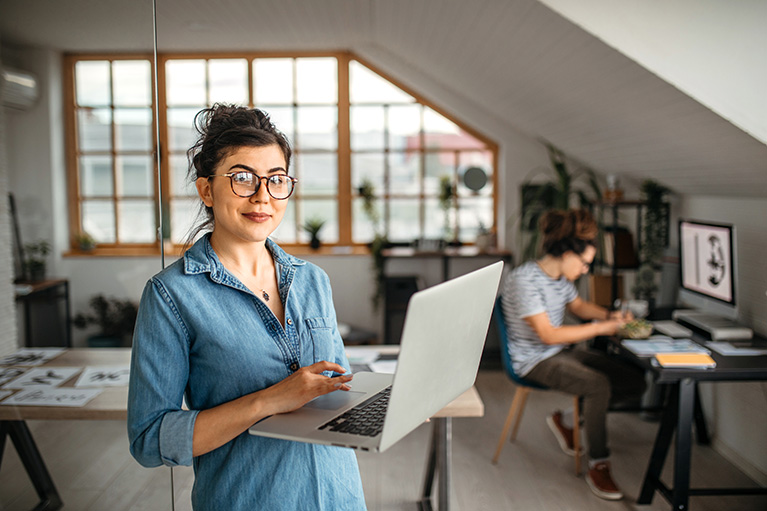 Femme entrepreneure souriante debout avec son ordinateur portable dans un espace de travail, tandis qu’un collègue travaille à son bureau en arrière-plan.