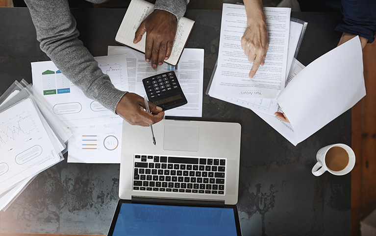 Photo des mains de deux personnes travaillant sur des documents et faisant des calculs. 