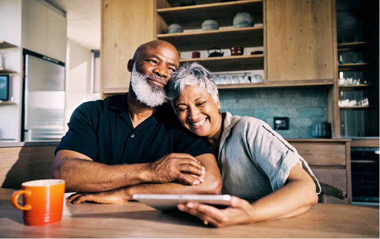 Un homme et une femme noir d'environ 60 ans assis sur leur table a diner souriant et lisant une une information sur une tablette.