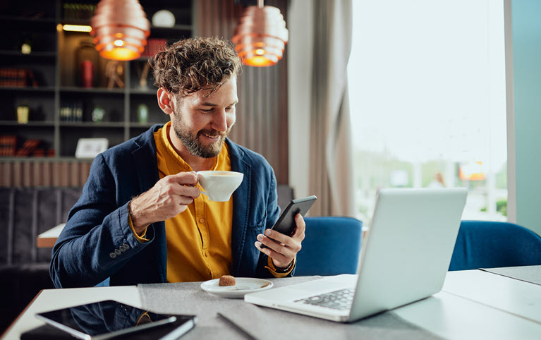 homme souriant assis avec une tasse en main devant son laptop, avec son cellulaire en main